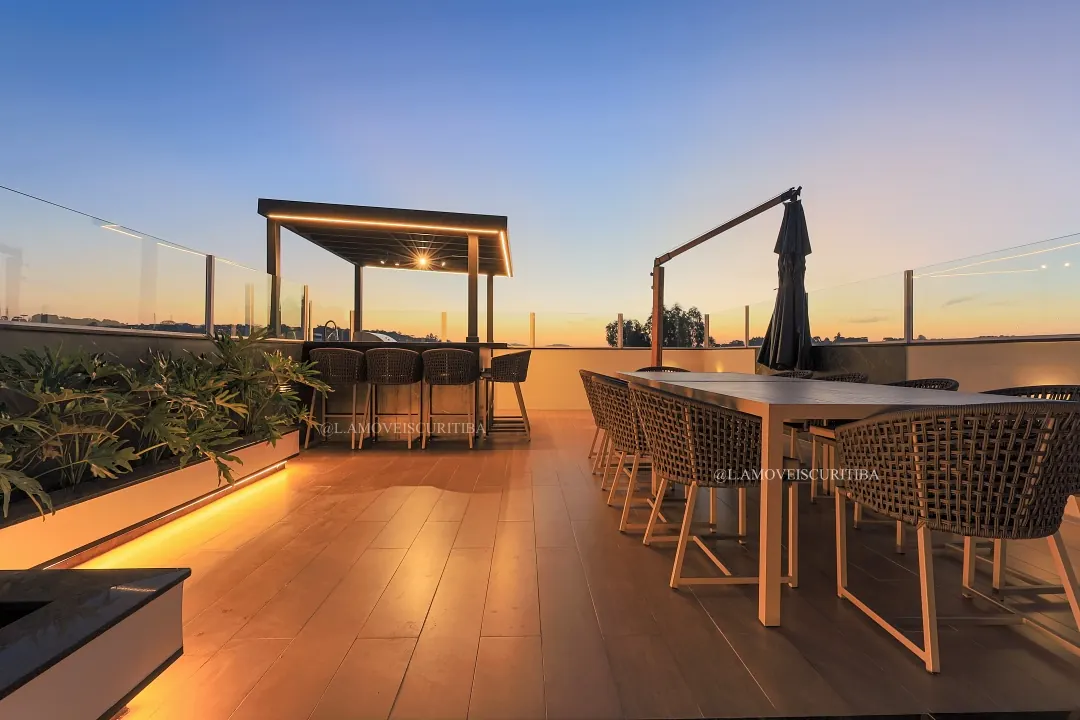 Deck com pergolado iluminado, bancada e mesa de jantar ao anoitecer