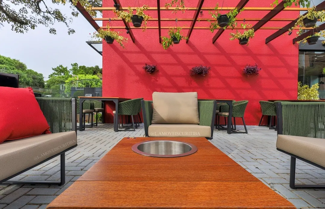 Lounge com mesa de centro em Cumaru e poltronas em corda náutica verde diante de parede vermelha sob o pergolado