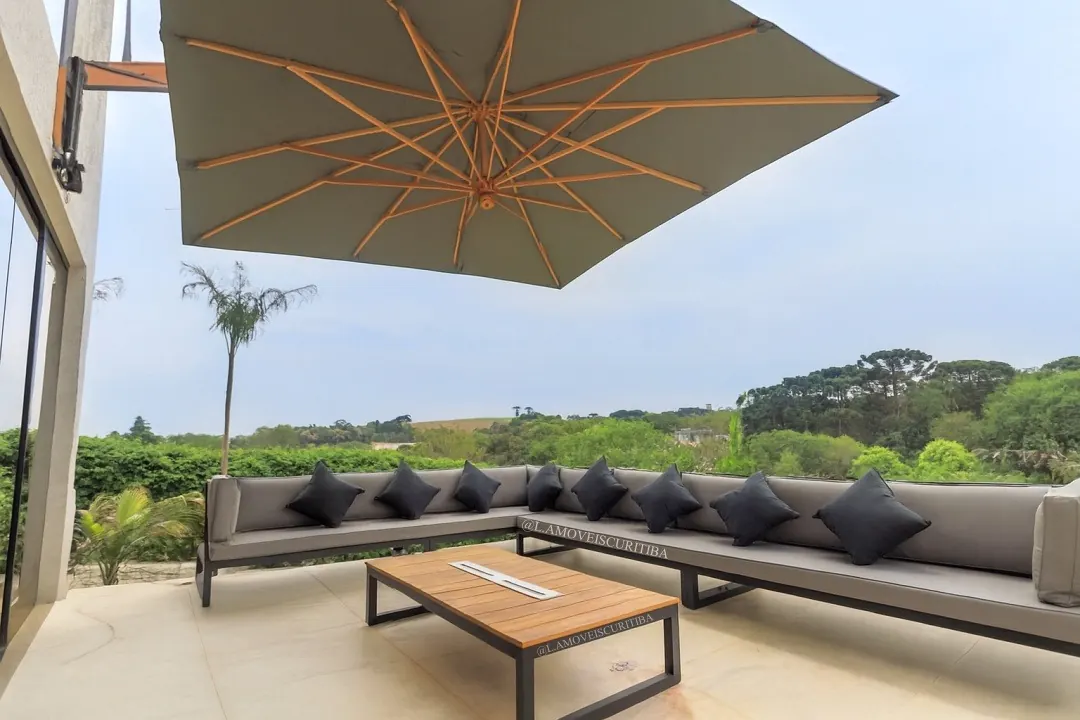 Sofá modular em L com almofadas cinza e pretas sob ombrelone de parede, em terraço com vista para a mata
