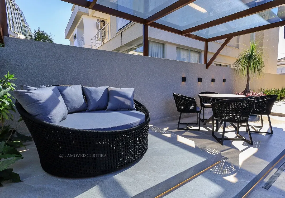 Chaise orbital preta com almofadas cinza sob pergolado envidraçado, ao lado de mesa de jantar